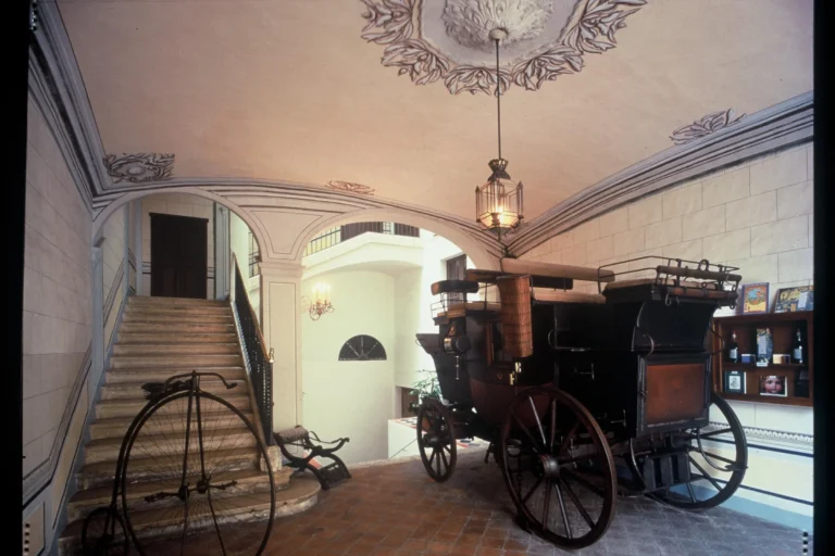 Intérieur du Museu Romàntic de Sitges avec une calèche historique, un vélo ancien et un escalier d'apparat au plafond décoré.