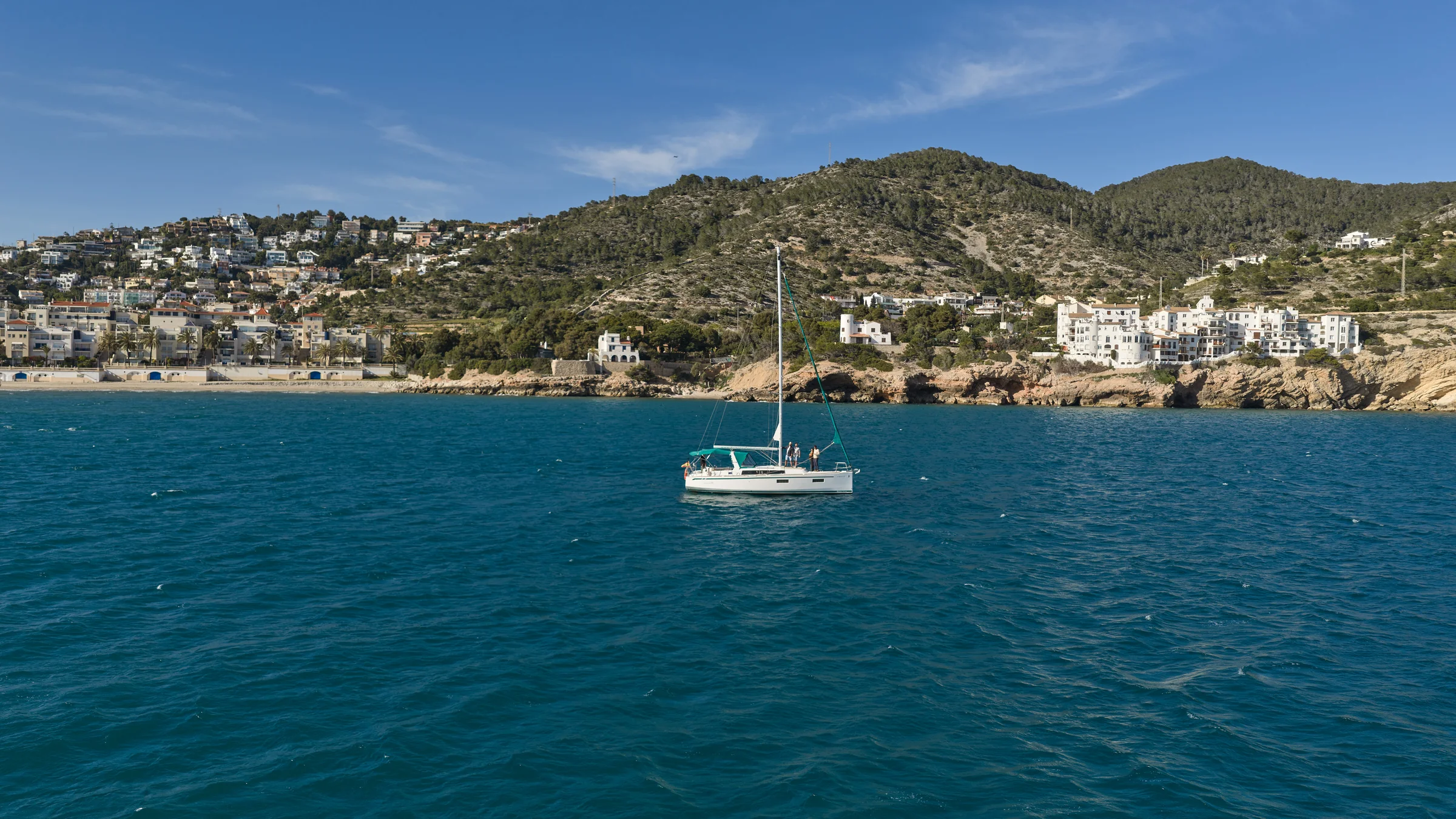 Velero navegando frente a la costa de Sitges con el mar Mediterráneo en primer término y las colinas y casas blancas de fondo.