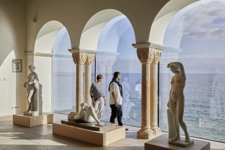 Deux visiteurs contemplent les sculptures noucentistes de Joan Rebull dans la salle du Mirador du Musée de Maricel de Sitges, avec la Méditerranée en arrière-plan.