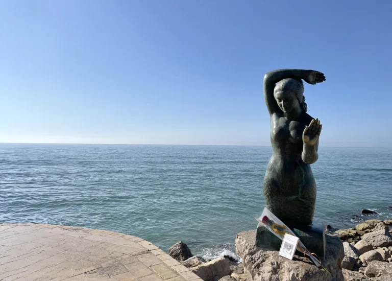 Estatua del Passeig Marítim de Sitges con una rosa y un libro a sus pies, en un día de Sant Jordi con el mar de fondo.